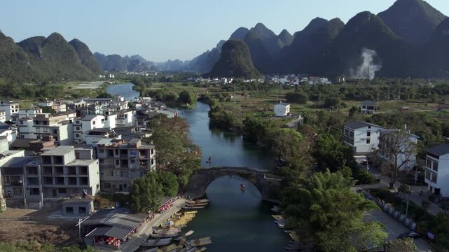 A rural riverside scene with an old stone arch bridge, flanked by buildings and karst mountains in the distance.