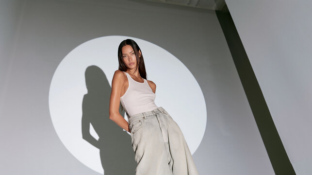 brunette woman in circular studio spotlight, young woman wearing white tank top and oversized denim jeans in studio, minimalist fashion portrait of female model posing in dramatic spotlight