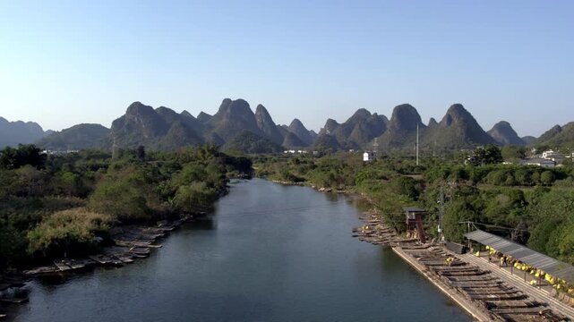 A calm river winds through lush greenery, framed by distinctive karst mountains under a clear blue sky, with boats moored along its banks.