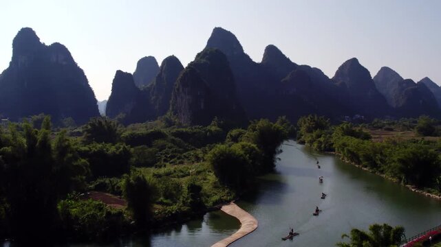 A scenic view of steep karst mountains flanking a winding river with small boats floating on calm water, surrounded by lush green vegetation.