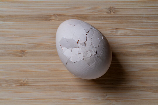Cracked chicken egg on wooden cutting board. Single white chicken egg with heavily cracked shell lies on light brown wooden textured surface, closeup, top view