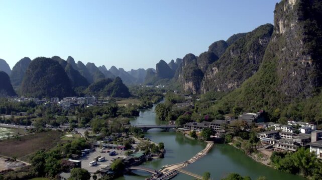A scenic landscape featuring karst mountains, a winding river, a village, and a bridge under a clear blue sky, showcasing natural and rural elements.