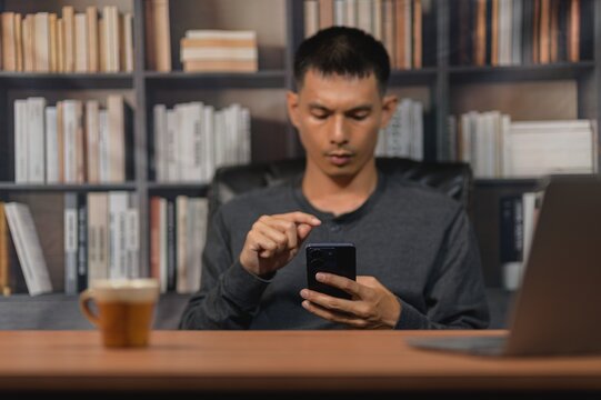 Young man using smartphone at desk in modern library with coffee and laptop