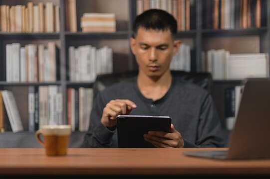 Young man using tablet device at desk with coffee and laptop in a library setting
