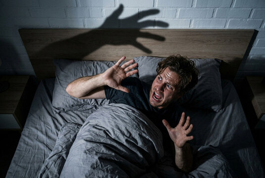 Scared man screaming in bed under terrifying monster hand shadow.