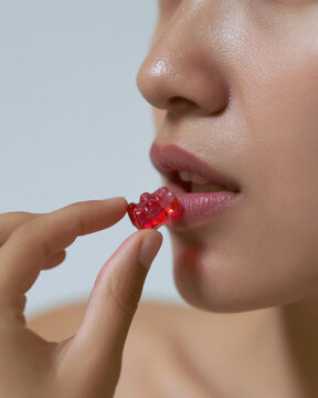 Melatonin gummy near mouth, close up of hand holding red gummy bear with lips and nose in soft focus, clean studio light grey background