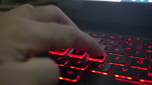 Close up of a gamer hand playing on a laptop computer keyboard with glowing red backlit keys, highlighting esports competition, action gaming, and modern entertainment technology.