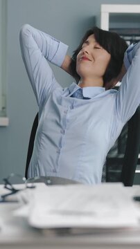 A woman sits at her desk in a busy office. She leans back in her chair, resting her arms behind her head and letting her mind wander away from work tasks.