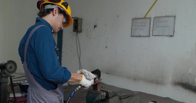 Factory worker using handheld grinder to cut metal workpiece in bench vise at workshop