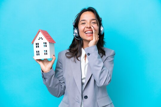 Real estate hispanic agent holding a toy house isolated on blue background shouting with mouth wide open
