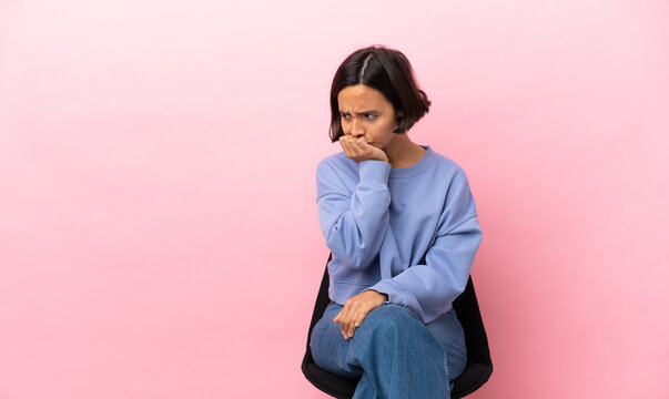 Young mixed race woman sitting on a chair isolated on pink background having doubts