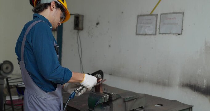 Factory worker using handheld grinder to cut metal workpiece in bench vise at workshop