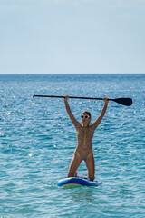 Fototapeta premium Paddleboarding, woman, ocean. Joyful woman kneeling on a paddleboard with paddle raised high, celebrating success during summer vacation, copy space