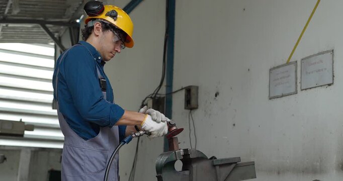 Factory worker using handheld grinder to cut metal workpiece in bench vise at workshop