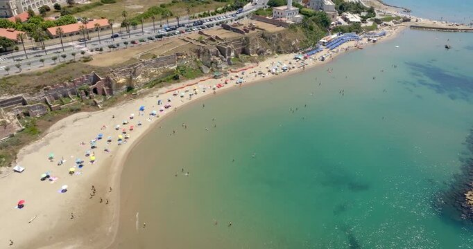 Aerial view of a sunny Mediterranean beach with sandy shorelines, ancient ruins along the coast, clear turquoise sea, breakwaters, and a nearby road. Panorama of Grotte di Nerone Beach in Anzio, Italy