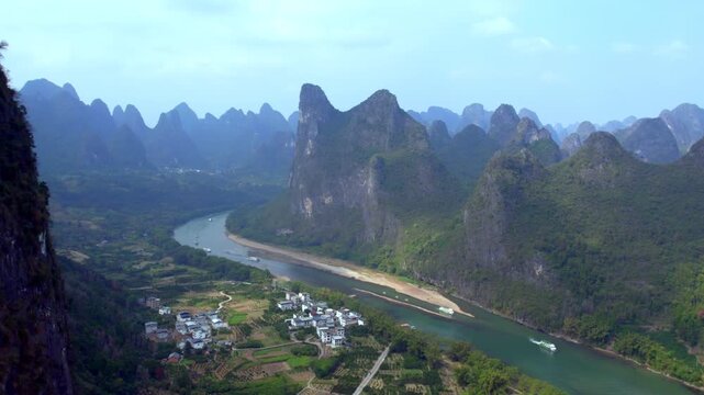 A high-angle view of a winding river meandering through lush karst mountains, with a small village and boats on the water.