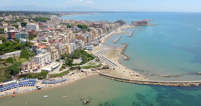 Aerial view of Anzio, Italy. It's a picturesque coastal city featuring sandy beaches, vibrant blue waters, a white lighthouse, and urban developments lining the scenic shore under a bright clear sky.