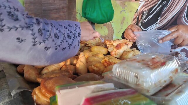 Indonesian buyer selecting various gorengan fried snacks at traditional market stall. Colorful assortment of fritters, pastries and jajanan pasar served on newspaper. Authentic street food shopping.