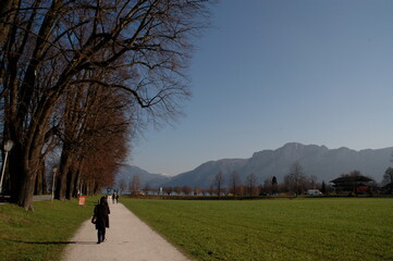 Obraz premium People walking along a rural pathway beside green fields and bare trees, with Alpine hills beneath a clear blue sky in Austria.