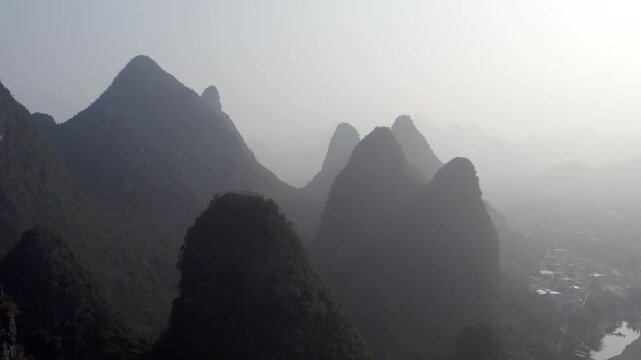 A misty landscape of steep limestone karst peaks rising above a winding river in a rural valley, with distant settlements partially obscured by haze.