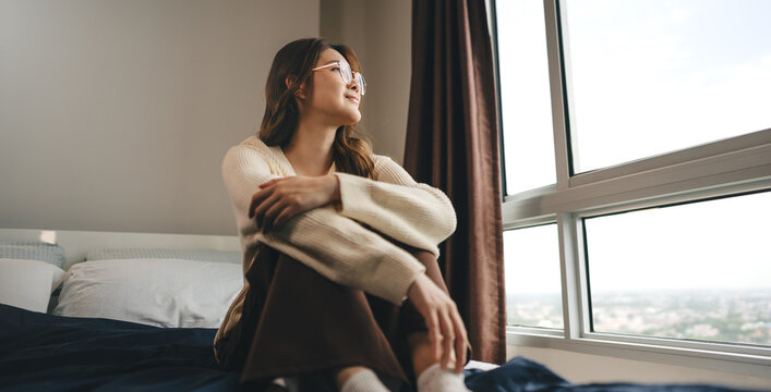 Happy young adult asian woman relax sitting on bed, wearing knitwear looking out window