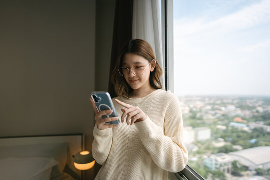 Young adult asian woman using smartphone ai chatbot technology standing near window in cozy home bedroom