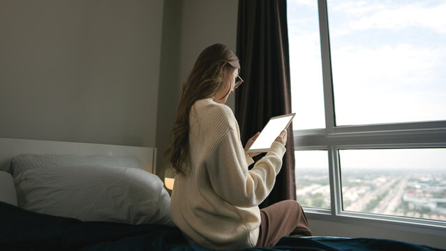 Behind sitting near window woman using digital tablet for ai chatbot technology  living in cozy home bedroom