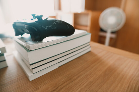 Black Xbox controller resting on a stack of white books on a wooden desk in a cozy home study, soft