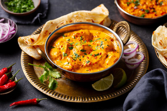 Butter chicken curry with flatbreads, onion, cilantro and lime on a brass tray. Dark background. Close up.