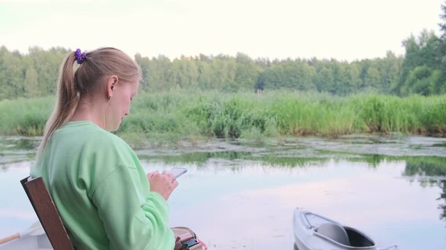 blonde woman types on phone by river ignoring peaceful nature. gadget addiction prevents enjoying beautiful landscape. digital distraction ruins outdoor relaxation and connection with environment.
