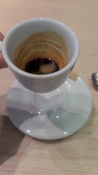 Close up of a person's hand picking up and swirling a nearly finished cup of traditional italian espresso coffee, revealing the remaining dark liquid and creamy residue on the sides