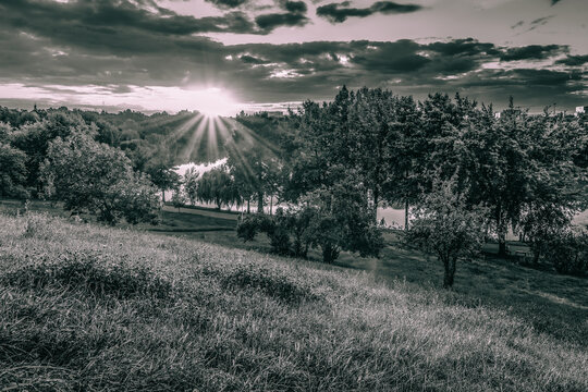 Black and white dramatic sunset with sun rays over a lake and meadow in a city park.