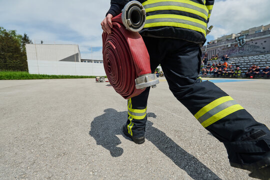 Firefighter Carrying Rolled Fire Hose During Outdoor Training Drill With Protective Gear And Team