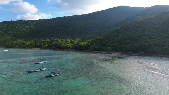 Aerial view of traditional fishing boats anchored in crystal clear tropical waters with lush green island coastline under blue sky