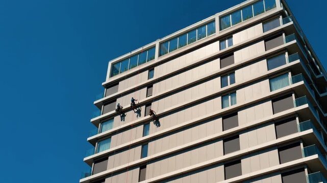 Industrial climbers cleaning windows on facade of modern building. Three workers hang on ropes against glass and concrete exterior under clear blue sky.