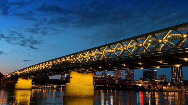 Stary most bridge illuminated over river at dusk in Bratislava. Green steel truss structure and city buildings reflect warm lights in water under evening sky.