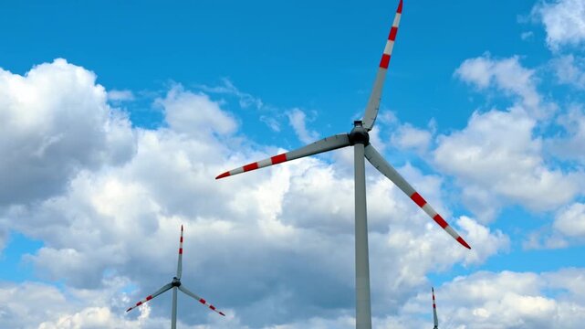 Low angle view of modern wind turbine against blue sky. Clean energy generator with rotating blades stands against white cumulus clouds in bright daylight.