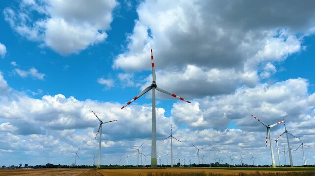 Wind turbine farm generating clean electricity under cloudy sky. Multiple white turbines, red tips, dry crop field, blue sky, cumulus clouds, bright daytime light.