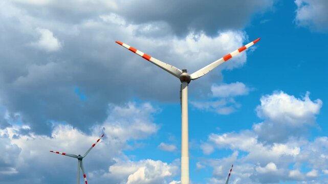 Upward view of wind turbine rotor with red striped blades. Large power generator with three rotor blades stands against blue sky filled with grey clouds.