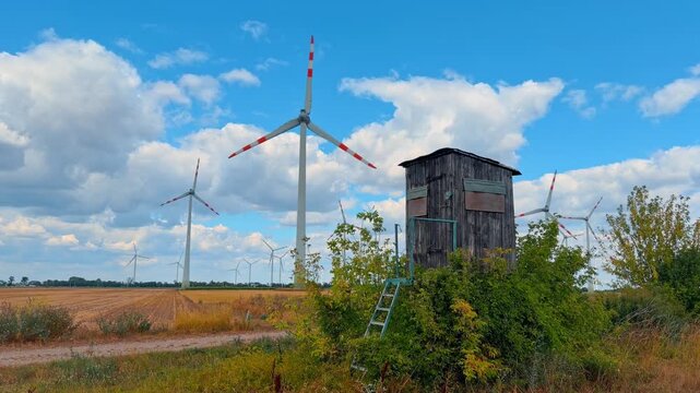 Wooden hunting stand near wind turbines in an open field under a blue sky. A timber observation tower rises above green vegetation with white generators visible in the distance.