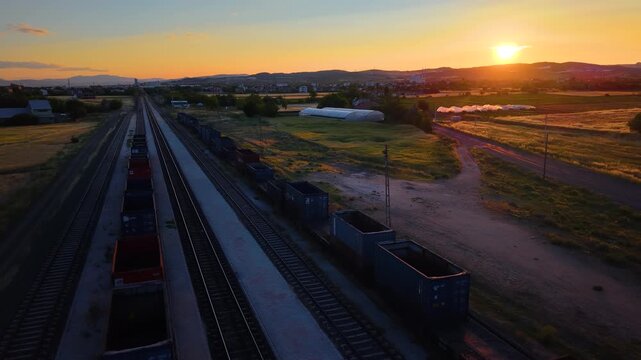 A smooth backward camera movement reveals freight train cars, greenhouse complexes and expansive farmland under the warm glow of the setting sun.
