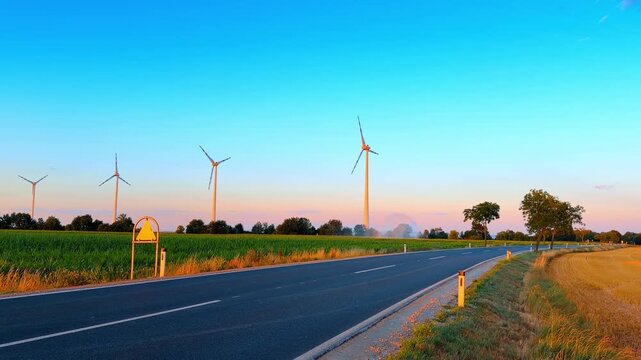 Asphalt road passing agricultural field and wind turbines. Tall white wind generators stand in a green farm field beside a curving rural highway under clear sky.