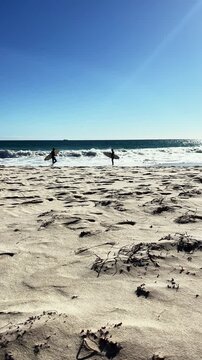 Vertical shot of two people carry surfboards across the sand toward the ocean to catch waves on a beautiful sunny day