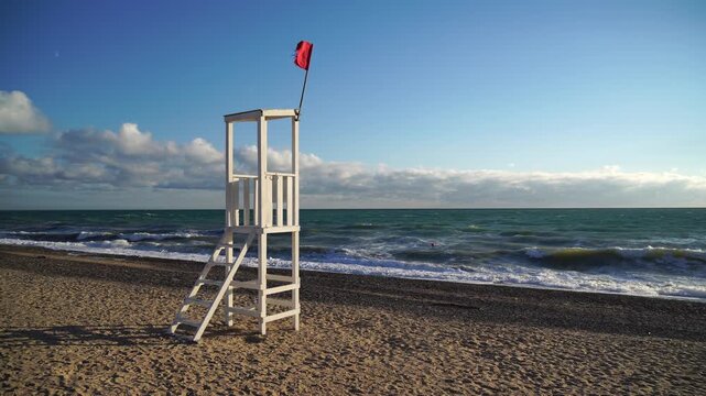 Lifeguard Tower Beach Red Flag Warning - A white lifeguard tower on a sandy beach with a red flag flying, indicating dangerous conditions for swimming.