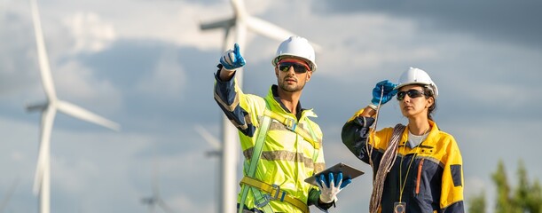 Fototapeta premium Workers teamwork inspecting wind turbine power generator sustainability performance inspection together at Renewable Energy Plant Man pointing and hold tablet with woman for check in wind farm