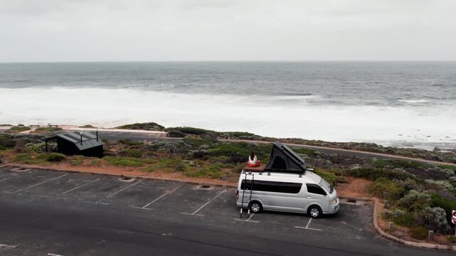 Woman sits on the roof rack of a parked camper van while looking at the large waves and gray ocean on an overcast day by the coast