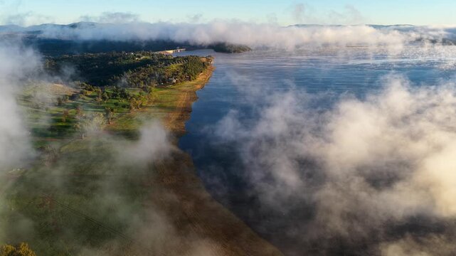 Aerial video of Lake Keepit Dam blanketed by morning fog in NSW, Australia. Low cloud drifts across the reservoir and surrounding landscape