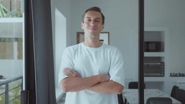 Success concept with young man standing with crossed arms and smiling confidently. Millennial male presents strong personality and self confidence in modern bright home environment.
