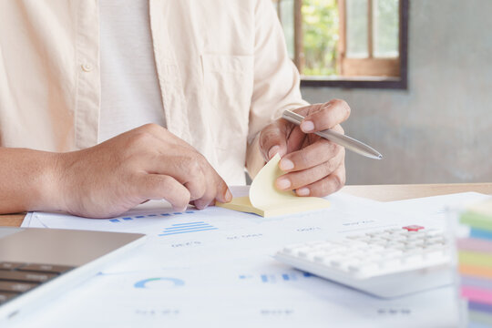 Close-up of business professional reviewing financial reports, writing notes on sticky paper, analyzing budget data, calculator and laptop in modern home office, financial planning, expense management
