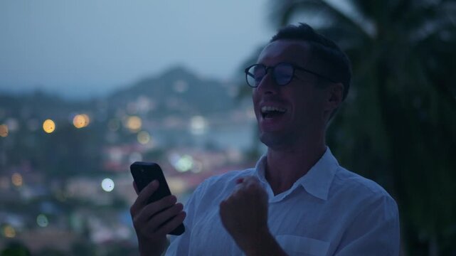 Winning moment, excited man checks smartphone and celebrates success with joyful yes gesture at dusk. Happy winner reacts to good news on mobile screen showing online victory luck and digital success